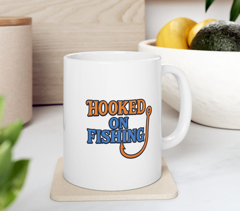 A white ceramic coffee mug displayed on table with bowl of fruit, featuring the phrase "Hooked on Fishing" in bold orange and blue text with a graphic of a fishing hook integrated into the design.