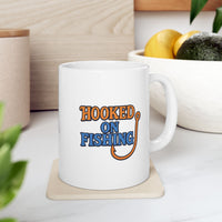 A white ceramic coffee mug displayed on table with bowl of fruit, featuring the phrase "Hooked on Fishing" in bold orange and blue text with a graphic of a fishing hook integrated into the design.