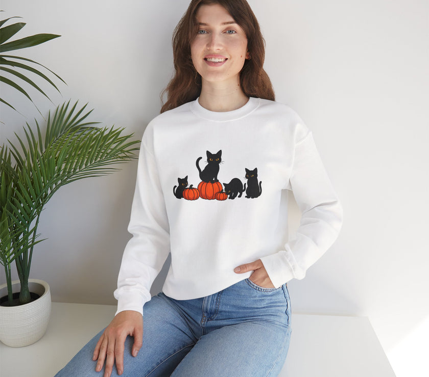 Woman wearing a white sweatshirt with black cat and orange pumpkin design, sitting on a white surface with a plant in the background.