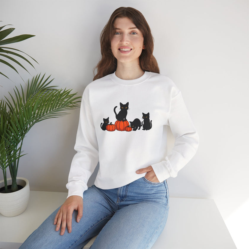 Woman wearing a white sweatshirt with black cat and orange pumpkin design, sitting on a white surface with a plant in the background.