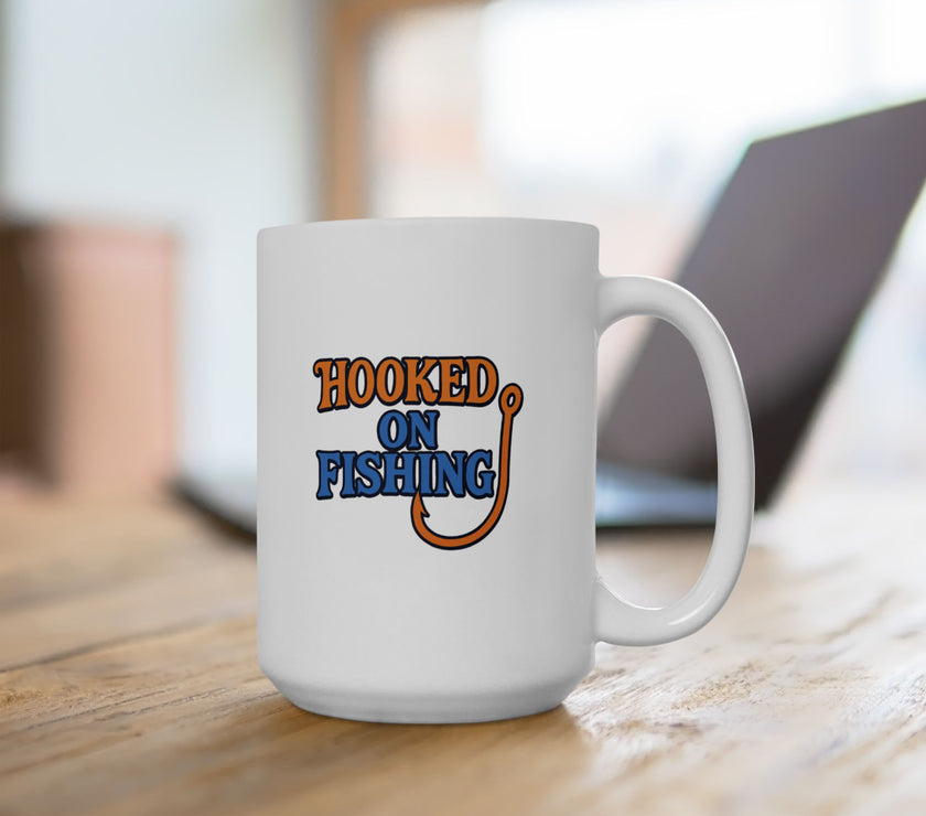 A white ceramic coffee mug displayed on a table with a laptop, featuring the phrase "Hooked on Fishing" in bold orange and blue text with a graphic of a fishing hook integrated into the design.
