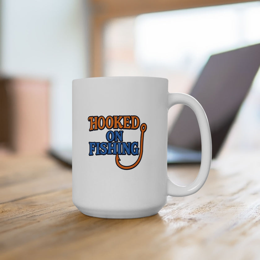 A white ceramic coffee mug displayed on a table with a laptop, featuring the phrase "Hooked on Fishing" in bold orange and blue text with a graphic of a fishing hook integrated into the design.