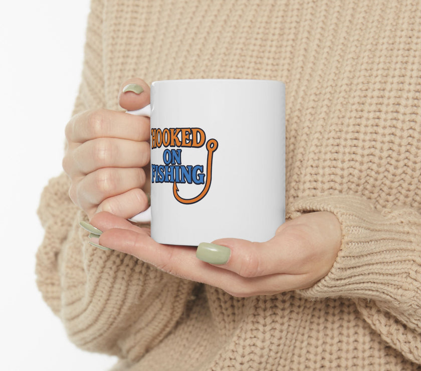 A white ceramic  featuring the phrase "Hooked on Fishing" in bold orange and blue text with a graphic of a fishing hook integrated into the design.