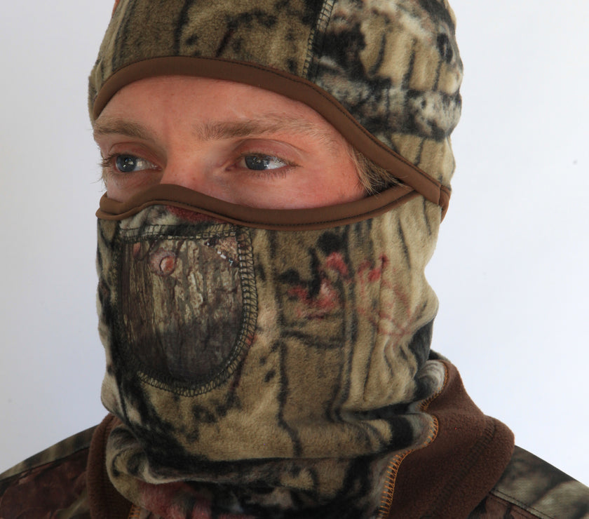 Person wearing a full-face camouflage fleece balaclava with pockets for disposable air-activated warmers on a white background