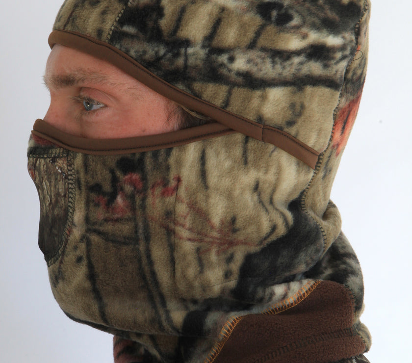 Person wearing a full-face camouflage fleece balaclava with pockets for disposable air-activated warmers on a white background