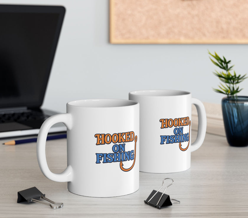 Two white ceramic coffee mug featuring the phrase "Hooked on Fishing" in bold orange and blue text with a graphic of a fishing hook integrated into the design.