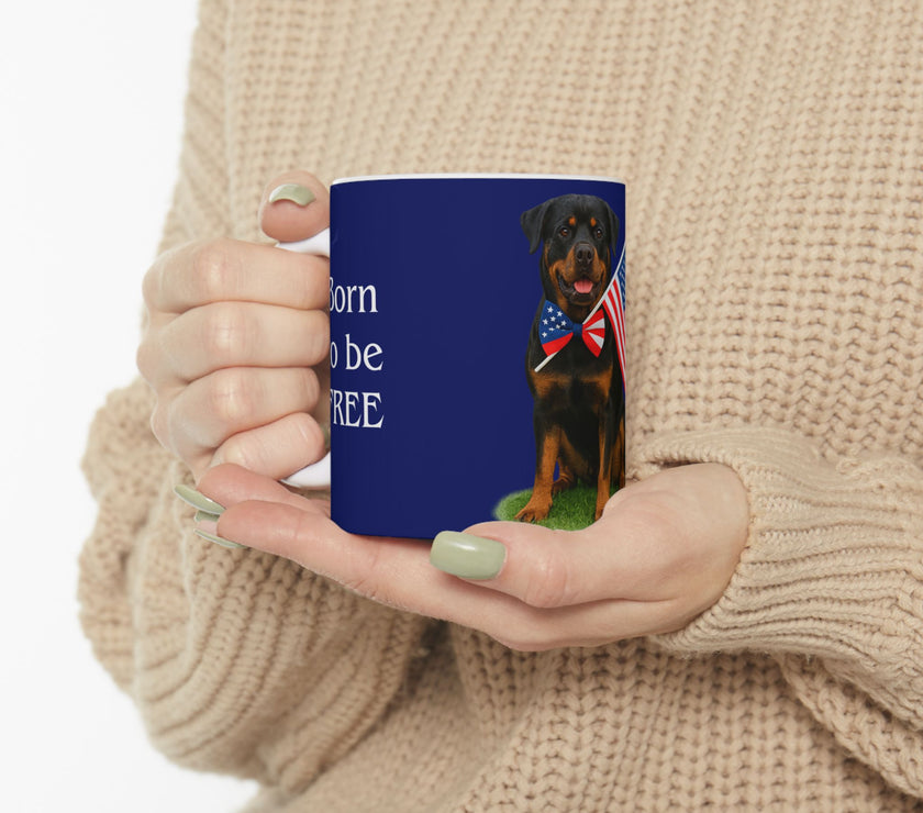 A ceramic mug featuring a Rottweiler sitting on grass, wearing a red, white, and blue bow tie, and holding an American flag against a dark blue background. with the message "Born to be Free"