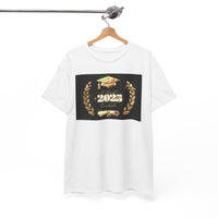 Light gray or white unisex T-shirt featuring a bold "Class of 2025 Graduates" design in gold, with a graduation cap, laurel wreath, and diploma printed on the front. T-shirt is on hanger