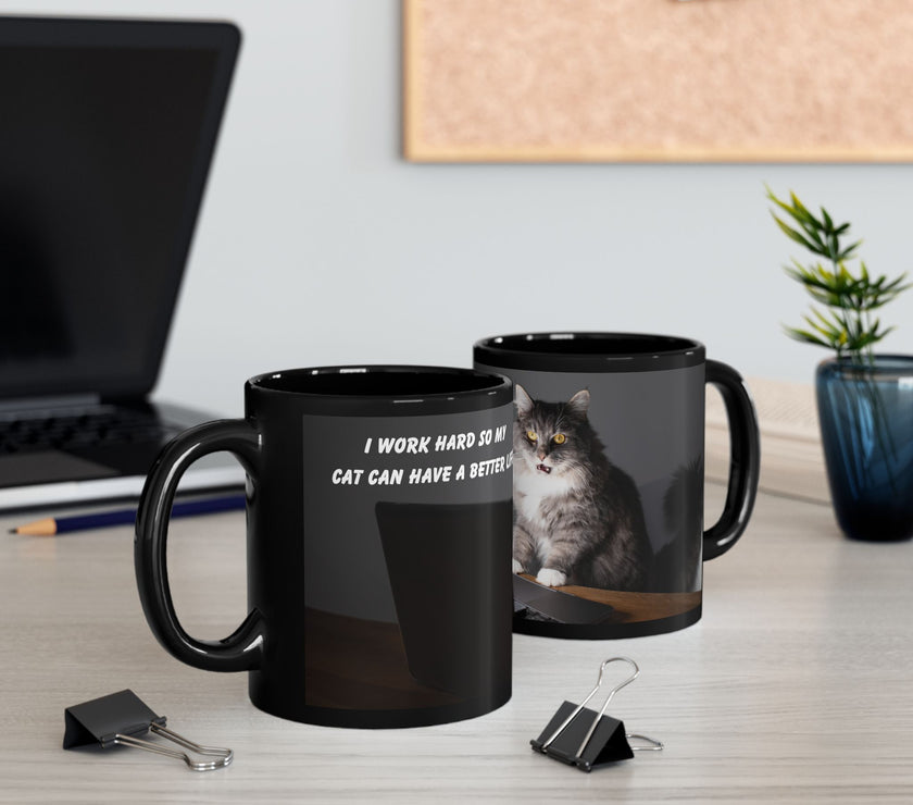 Black ceramic mug featuring a cartoon-style image of a cat sitting at a computer with the funny quote “I work hard so my cat can have a better life.”