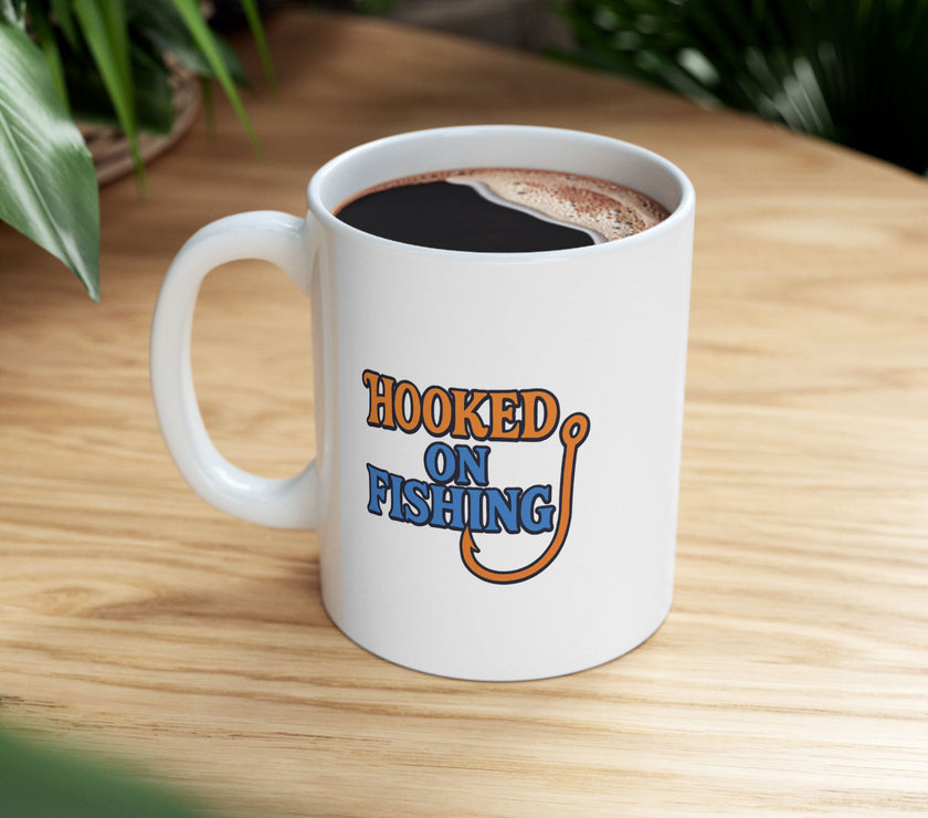A white ceramic coffee mug displayed on a table top, featuring the phrase "Hooked on Fishing" in bold orange and blue text with a graphic of a fishing hook integrated into the design.
