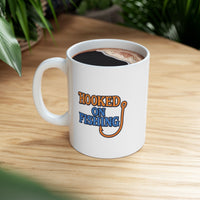 A white ceramic coffee mug displayed on a table top, featuring the phrase "Hooked on Fishing" in bold orange and blue text with a graphic of a fishing hook integrated into the design.