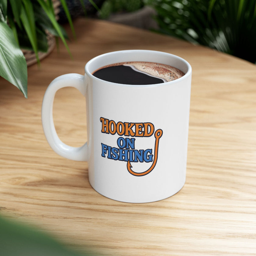 A white ceramic coffee mug displayed on a table top, featuring the phrase "Hooked on Fishing" in bold orange and blue text with a graphic of a fishing hook integrated into the design.