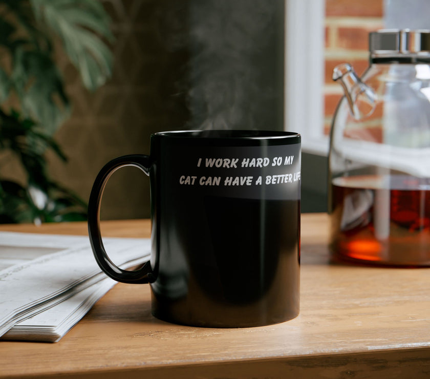 Black ceramic mug featuring a cartoon-style image of a cat sitting at a computer with the funny quote “I work hard so my cat can have a better life.”