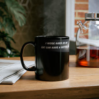 Black ceramic mug featuring a cartoon-style image of a cat sitting at a computer with the funny quote “I work hard so my cat can have a better life.”