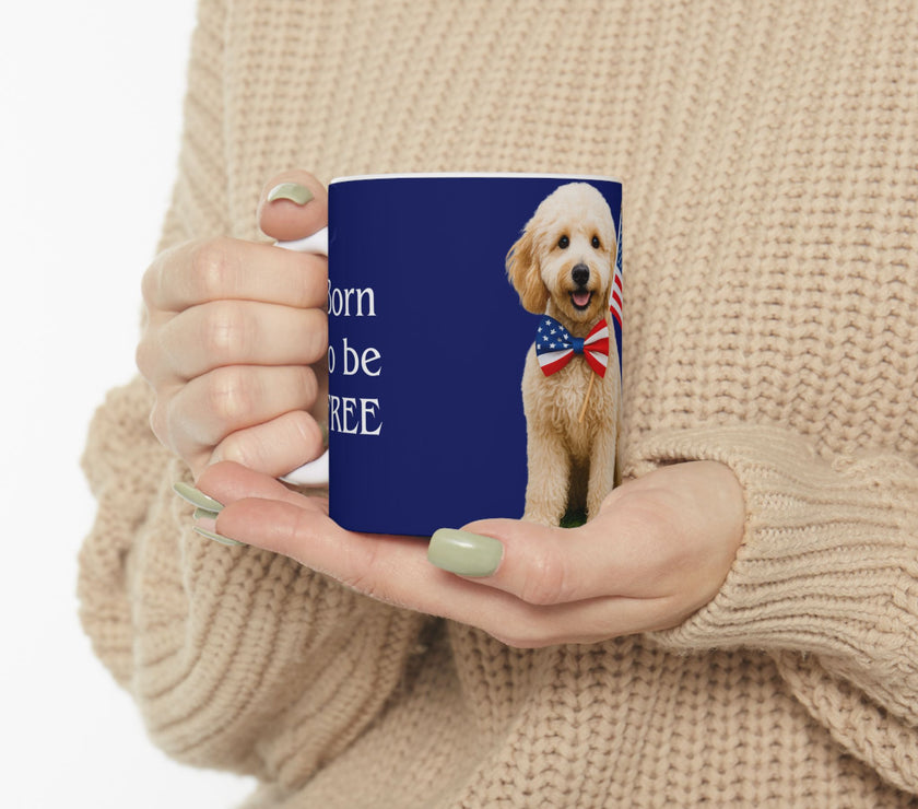 A ceramic mug featuring aGoldendoodle sitting on grass, wearing a red, white, and blue bow tie, and holding an American flag against a dark blue background. with the message "Born to be Free"
