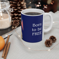 A ceramic mug featuring aGoldendoodle sitting on grass, wearing a red, white, and blue bow tie, and holding an American flag against a dark blue background. with the message "Born to be Free" side view