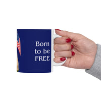 A ceramic mug featuring aGoldendoodle sitting on grass, wearing a red, white, and blue bow tie, and holding an American flag against a dark blue background. with the message "Born to be Free"