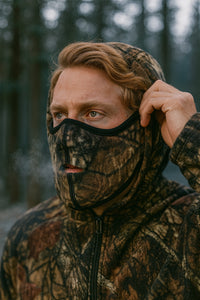 Person wearing a camouflage hood and mask in a forest setting
