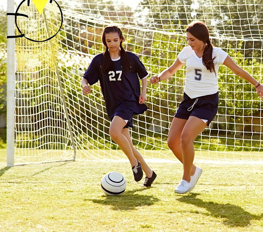 Two young girls playing soccer on a grass field with a goalpost in the background.