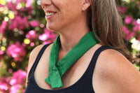 A woman wearing a green cooling neck wrap tied around her neck, standing outdoors with pink flowers blurred in the background.