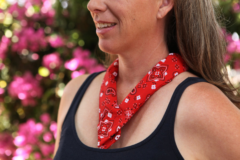 A woman wearing a red bandana cooling neck wrap tied around her neck, standing outdoors with pink flowers blurred in the background.