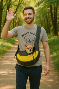 A smiling man walks along a wooded trail wearing a gray "River’s Corner" T-shirt and carrying a small dog in a yellow and gray sling pet carrier across his chest.