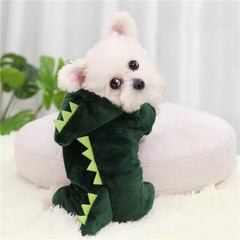 Small white dog wearing a green dinosaur costume on a white cushion with a plant in the background