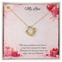 Gold love knot necklace with a sparkling center stone displayed in a red gift box on a soft Valentine-themed background with hearts and romantic message card.