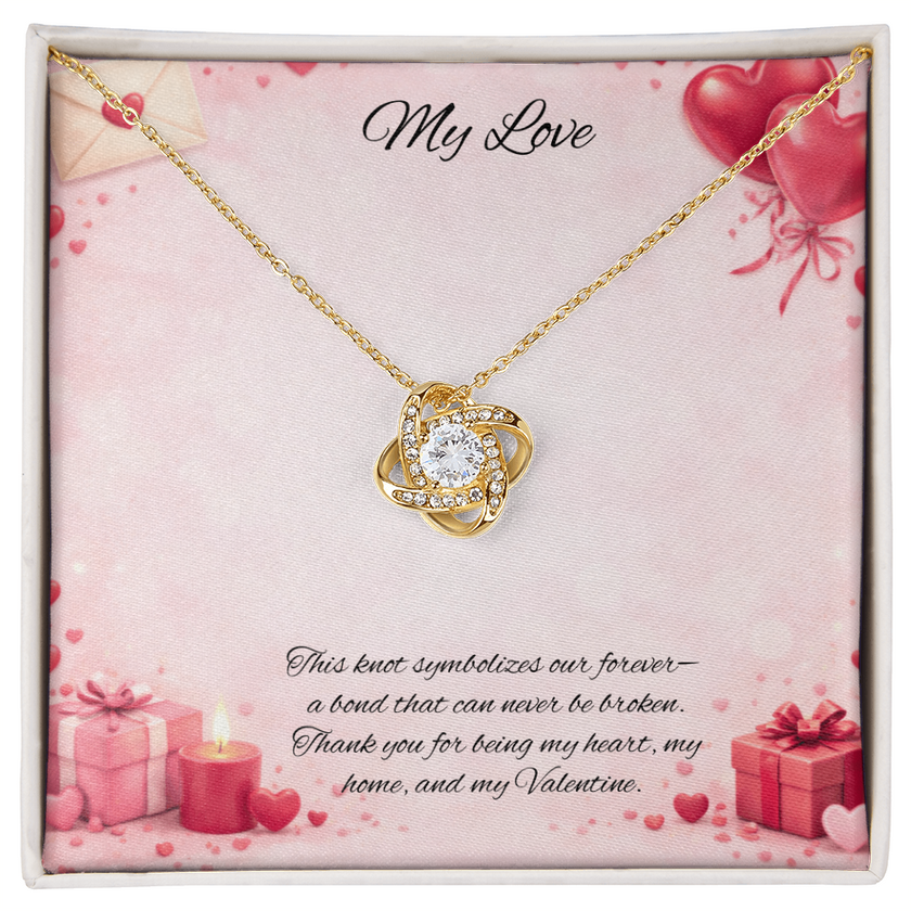 Gold love knot necklace with a sparkling center stone displayed in a red gift box on a soft Valentine-themed background with hearts and romantic message card.