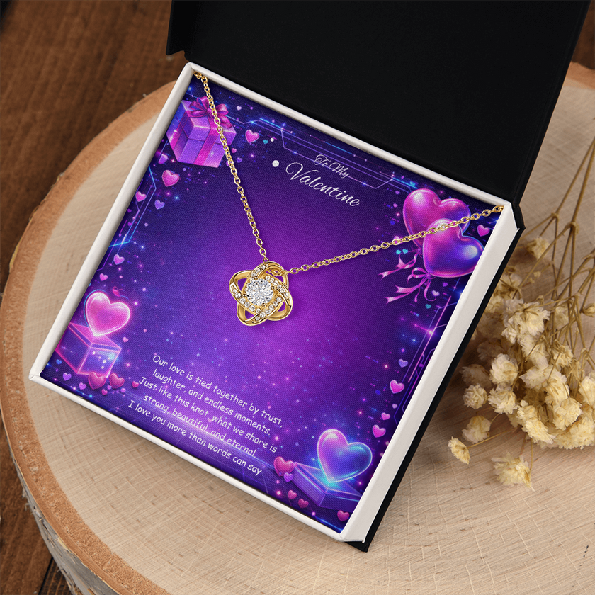 Gold love knot necklace with a sparkling center stone displayed on a purple Valentine’s Day background with glowing hearts, symbolizing eternal love and romantic connection.