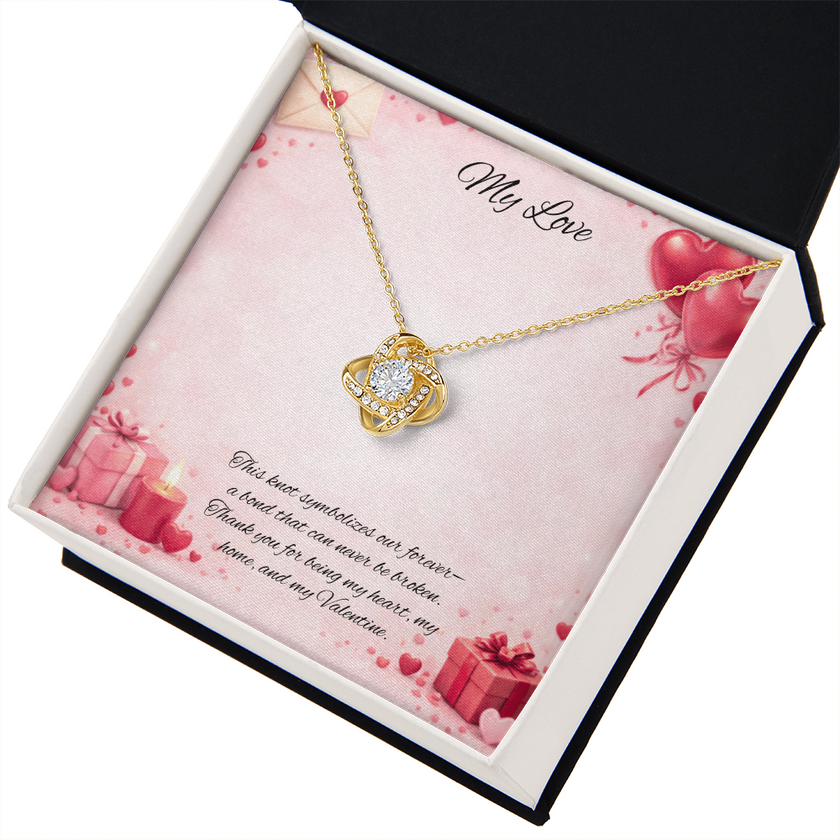 Gold love knot necklace with a sparkling center stone displayed in a  gift box on a soft Valentine-themed background with hearts and romantic message card.