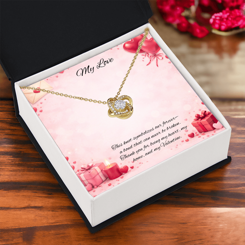 Gold love knot necklace with a sparkling center stone displayed in a  gift box on a soft Valentine-themed background with hearts and romantic message card.