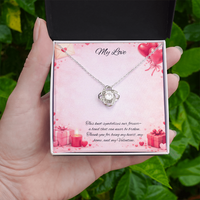 Silver love knot necklace with a sparkling center stone displayed in a gift box on a soft Valentine-themed background with hearts and romantic message card.