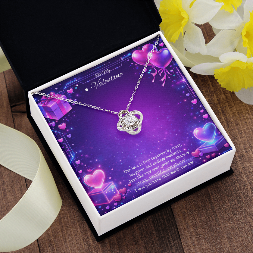 Silver love knot necklace with a sparkling center stone displayed on a purple Valentine’s Day background with glowing hearts, symbolizing eternal love and romantic connection.