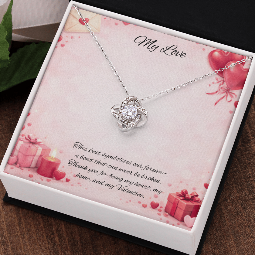 Silver love knot necklace with a sparkling center stone displayed in a gift box on a soft Valentine-themed background with hearts and romantic message card.