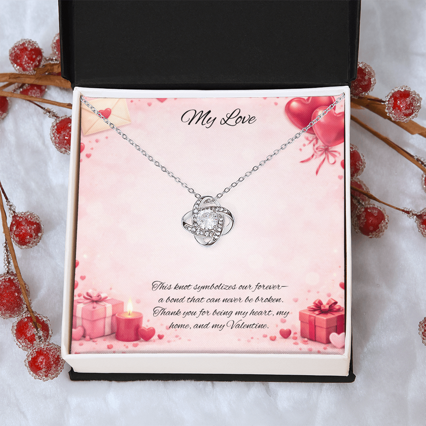 Silver love knot necklace with a sparkling center stone displayed in a  gift box on a soft Valentine-themed background with hearts and romantic message card.