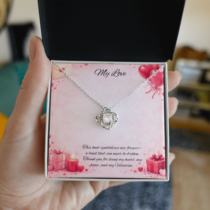 Silver love knot necklace with a sparkling center stone displayed in a gift box on a soft Valentine-themed background with hearts and romantic message card.