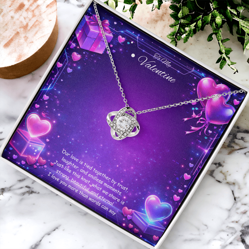 Silver love knot necklace with a sparkling center stone displayed on a purple Valentine’s Day background with glowing hearts, symbolizing eternal love and romantic connection.