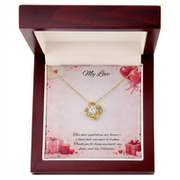 Gold love knot necklace with a sparkling center stone displayed in a red gift box on a soft Valentine-themed background with hearts and romantic message card.
