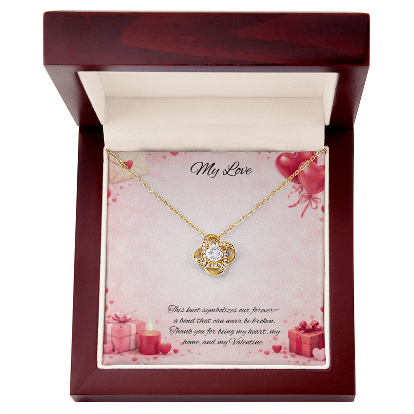 Gold love knot necklace with a sparkling center stone displayed in a red gift box on a soft Valentine-themed background with hearts and romantic message card.