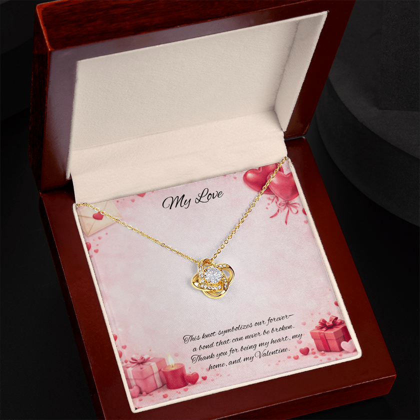 Gold love knot necklace with a sparkling center stone displayed in a red gift box on a soft Valentine-themed background with hearts and romantic message card.