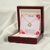 Gold love knot necklace with a sparkling center stone displayed in a red gift box on a soft Valentine-themed background with hearts and romantic message card.