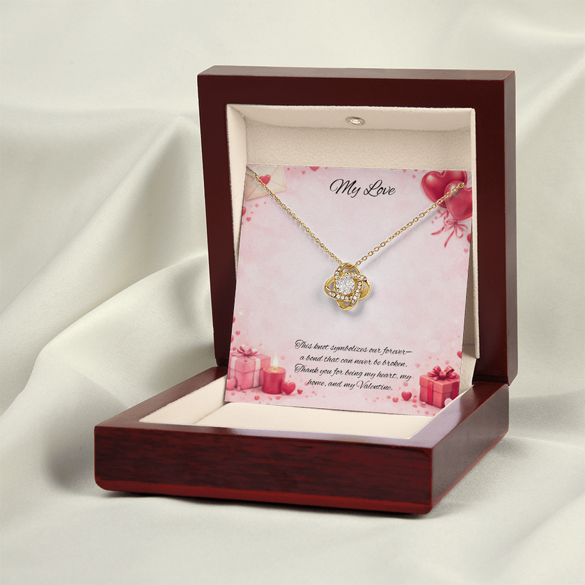 Gold love knot necklace with a sparkling center stone displayed in a red gift box on a soft Valentine-themed background with hearts and romantic message card.