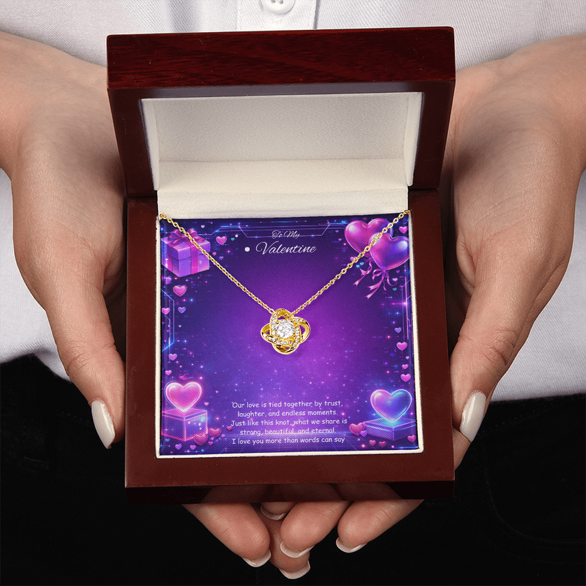 Gold love knot necklace with a sparkling center stone displayed on a purple Valentine’s Day background with glowing hearts, symbolizing eternal love and romantic connection.