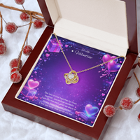 Gold love knot necklace with a sparkling center stone displayed on a purple Valentine’s Day background with glowing hearts, symbolizing eternal love and romantic connection.