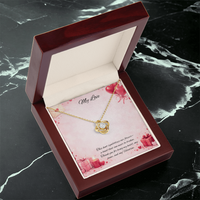 Gold love knot necklace with a sparkling center stone displayed in a red gift box on a soft Valentine-themed background with hearts and romantic message card.