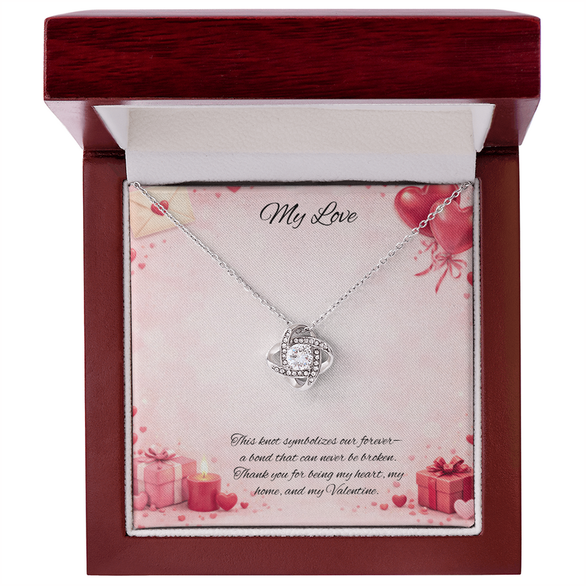Silver love knot necklace with a sparkling center stone displayed in a red gift box on a soft Valentine-themed background with hearts and romantic message card.