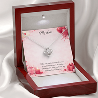 Silver love knot necklace with a sparkling center stone displayed in a red gift box on a soft Valentine-themed background with hearts and romantic message card.