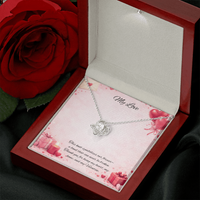 Silver love knot necklace with a sparkling center stone displayed in a red gift box on a soft Valentine-themed background with hearts and romantic message card.