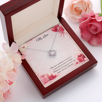 Silver love knot necklace with a sparkling center stone displayed in a red gift box on a soft Valentine-themed background with hearts and romantic message card.